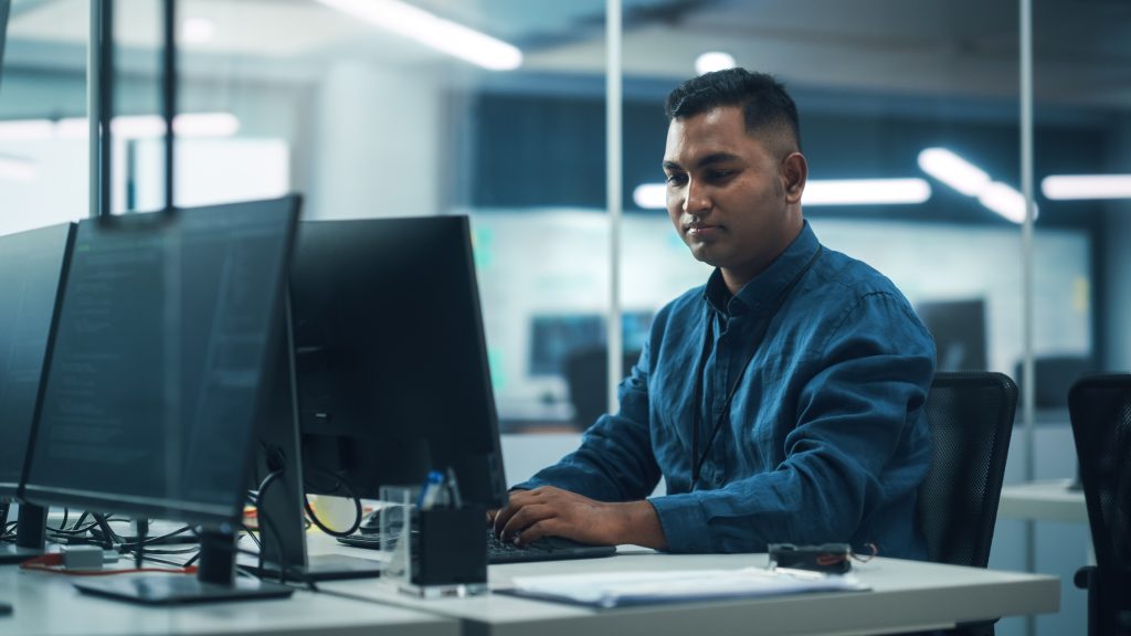 In Diverse Office: Portrait of Handsome Indian Man Working on Desktop Computer. Professional Programmer Creates Innovative Software, Modern App Design. Stylish Multi-Ethnic Authentic Workplace